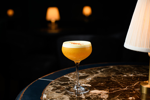 Orange alcoholic cocktail with a white scum decorated with orange zest on the marble table under the light