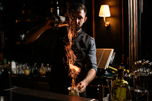 Bartender pouring a transparent alcoholic drink from the one steel cup to another making a fire on the bar counter