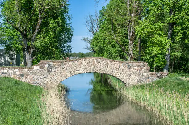 Old stone single arch bridge, Latvia. Famous ancient stone arch single track road bridge in the forest. View of small river and trees reflection in the water.