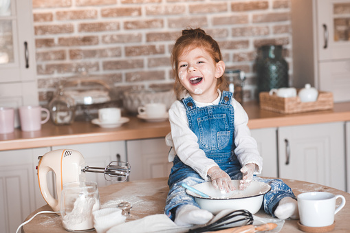 Laughing baby girl 3-4 year old making pastry sitting on table in kitchen closeup. Looking at camera. Childhood.