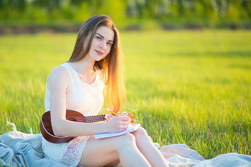 A girl on a sunny day sits on a plaid, plays on a ukulele, composes a song and writes the lyrics to a notebook in a field with green fresh grass