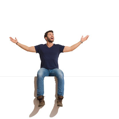 Confident and happy young man in boots, jeans and blue shirt is sitting on a top with arms outstretched, looking up and shouting. Full length studio shot isolated on white.