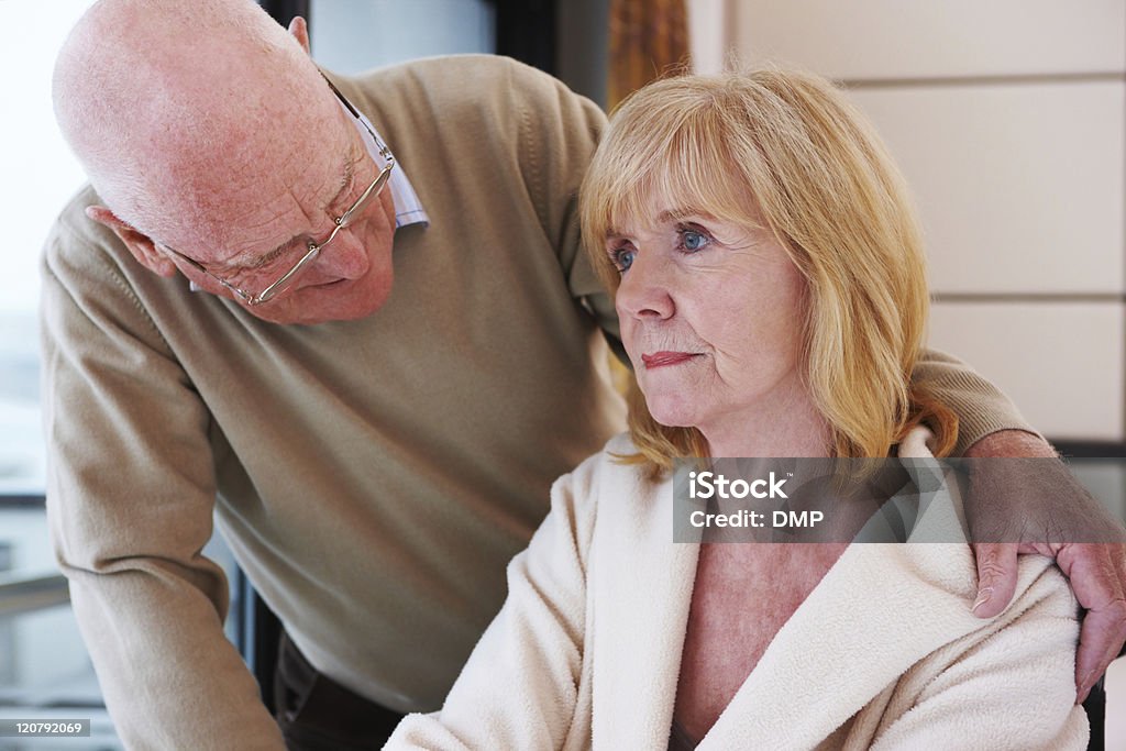 Senior Man Offering Comfort Senior man affectionately stands with an arm around his wife at the hospital. She is sitting with a blank expression on her face. Horizontal shot. Dementia Stock Photo