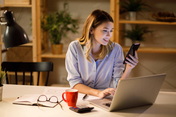 istock Woman holding smartphone and working on laptop at night 1207924575