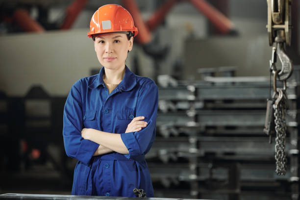 junge selbstbewusste frau in hardhat, handschuhen und blauer arbeitskleidung, die sie anschaut - manager foreman warehouse arms crossed stock-fotos und bilder