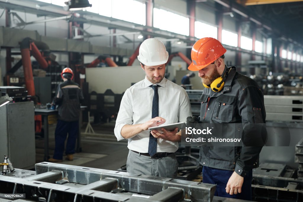 Young master in hardhat and bearded engineer discussing technical sketch Young master in hardhat and bearded engineer discussing technical sketch on display of tablet in factory workshop Industry Stock Photo