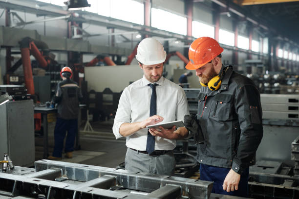 Young master in hardhat and bearded engineer discussing technical sketch Young master in hardhat and bearded engineer discussing technical sketch on display of tablet in factory workshop quality control photos stock pictures, royalty-free photos & images