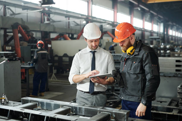 Young master in hardhat and bearded engineer discussing technical sketch Young master in hardhat and bearded engineer discussing technical sketch on display of tablet in factory workshop foreman stock pictures, royalty-free photos & images