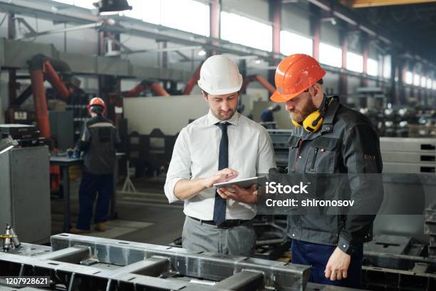 Young Master In Hardhat And Bearded Engineer Discussing Technical Sketch Stock Photo - Download Image Now