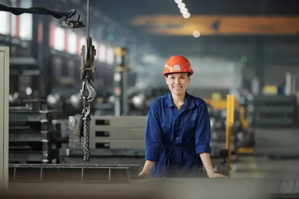 Cheerful young woman in workwear and protective helmet looking at you while working in modern industrial plant