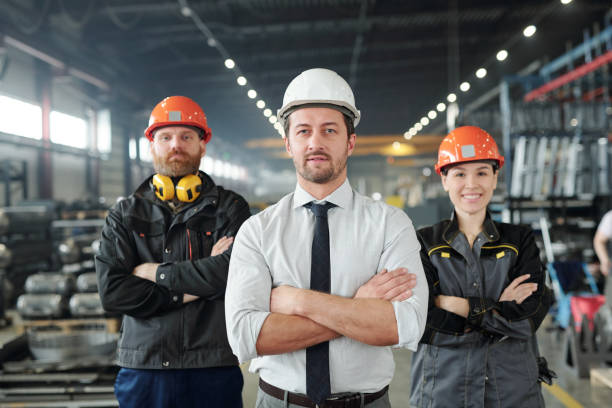 joven capataz en ropa formal y hardhat y sus dos subordinados - manager foreman warehouse arms crossed fotografías e imágenes de stock