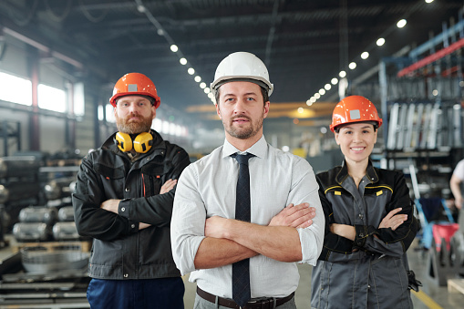 Young foreman in formalwear and hardhat and his two confident subordinates in workwear standing in front of camera
