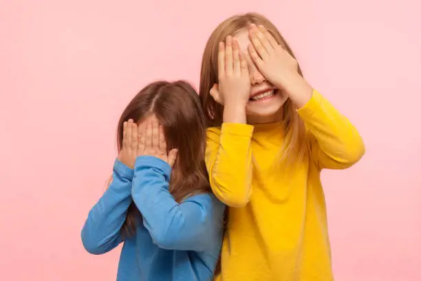 Portrait of cheerful joyful carefree little girls covering their faces with arms and sincerely smiling, laughing, having fun and playing peek a boo game. indoor studio shot isolated on pink background
