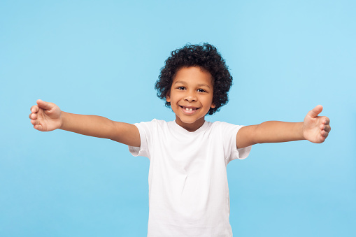 Come here, free hugs! Portrait of lovely good-natured little boy with curly hair in white T-shirt smiling excitedly and holding hands wide open to embrace, greeting. indoor studio shot blue background
