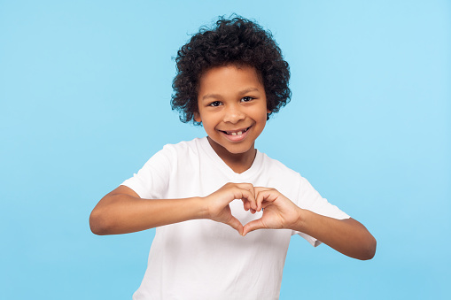 Portrait of smiling cheerful little boy with curly hair in white T-shirt showing heart shape with fingers, expressing innocent childhood love, affection. indoor studio shot isolated on blue background