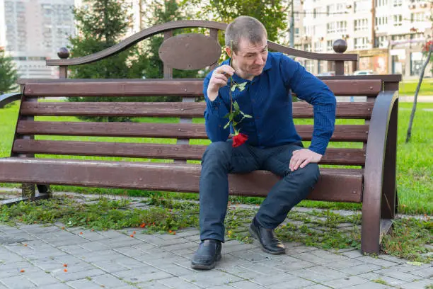 Photo of A young man sitting on a bench with a rose in his hands looks with contempt at passers-by