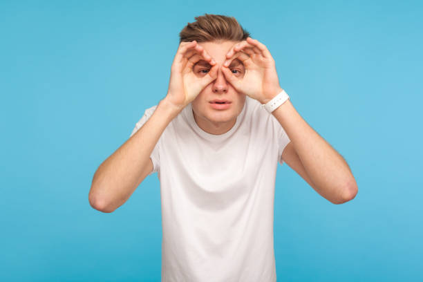 retrato de homem curioso em camiseta branca casual olhando através dos dedos em gestos binóculos, observando distante com olhar atento - distant sign - fotografias e filmes do acervo