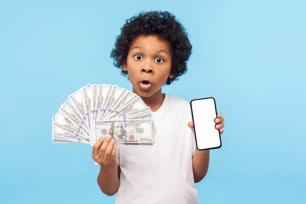 adorable cute surprised little boy with curly hair in t-shirt holding lots of money and cell phone - paying children only retail childhood imagens e fotografias de stock