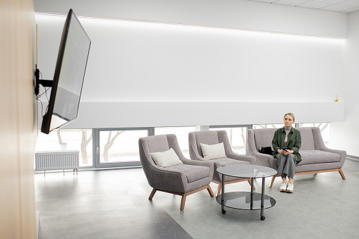 Young casual woman sitting on couch inside contemporary medical center and watching commercial on display while waiting for her turn