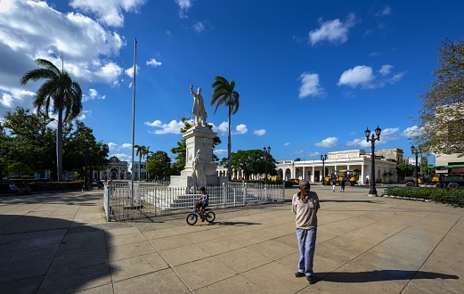 Cienfuegos, Cuba, December 3, 2017: A elderly man walks and a boy rides bicycle in the José Martí Park in the city center. The park bears the name of the Cuban revolutionary philosopher and political theorist José Martí whose statue is seen in the center of the square. In the background is the Arch of Triumph dedicated to Cuban independence. The historic center of Cienfuegos is listed as UNESCO World Heritage Site.