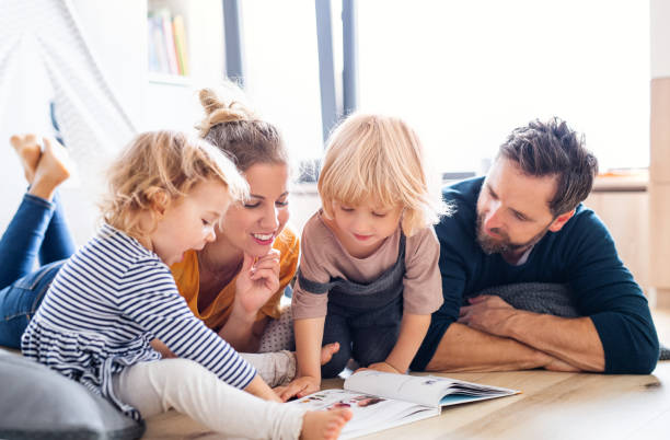 família jovem com duas crianças pequenas dentro de casa no quarto lendo um livro. - family reading book child - fotografias e filmes do acervo