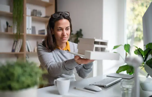 Architect with model of a house sitting at the desk indoors in office, working.