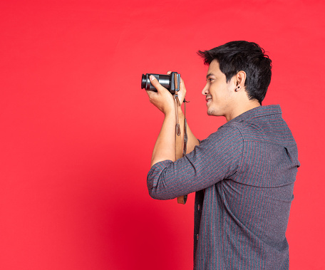 Side view of hipster in casual fashion shirt taking photo with dslr camera. half body portrait isolated on red studio background.
