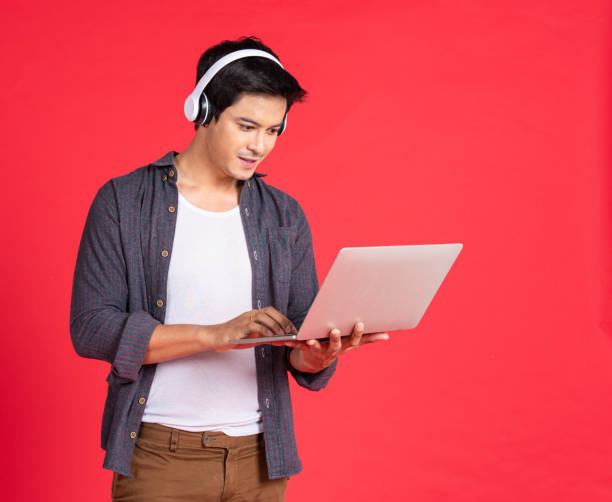 giovane hipster bel ragazzo asiatico con auricolari wireless in testa che giocano su computer portatile mentre si trova isolato su un adolescente rosso che ascolta musica - dj stand foto e immagini stock