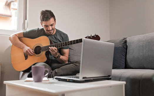 Young man playing acoustic guitar while sitting on a sofa in his living room.