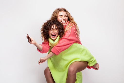 Two cheerful beautiful women in bright colorful sweaters having fun together against white background