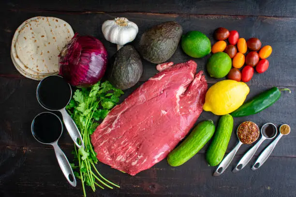 Raw flank steak with fresh vegetables and seasonings on a dark wood background