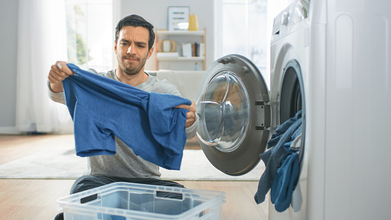 Handsome Confused Young Man in Grey Jeans and Coat Sits in Front of a Washing Machine at Home. He Loads the Washer with Dirty Laundry. Bright and Spacious Living Room with Modern Interior.