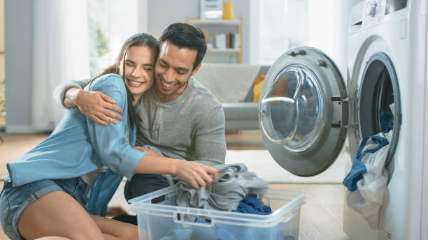 schönes junges paar sitzt neben einer waschmaschine zu hause. sie lachen und umarmen, während sie die waschmaschine mit schmutziger wäsche laden. helles und geräumiges wohnzimmer mit modernem interieur. - laundry husband housework men stock-fotos und bilder