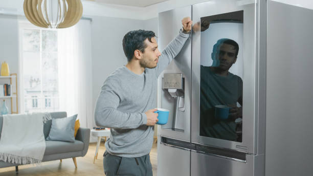 istock Handsome Young Man is Standing Next to a Refrigerator While Drinking His Morning Coffee. He is Checking a To Do List on a Smart Fridge at Home. Kitchen is Bright and Cozy. 1208032991