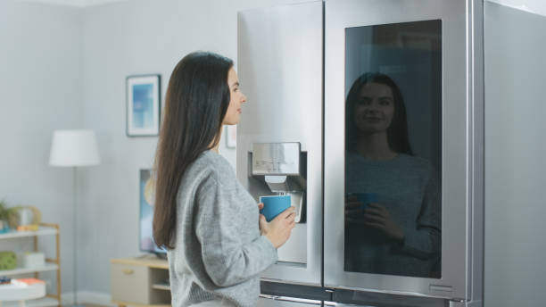 istock Beautiful Young Girl Stands Next to a Refrigerator While Drinking Her Morning Coffee. She is Checking the Weather Forecast and a To Do List on a Smart Fridge at Home. Kitchen is Bright and Cozy. 1208032997