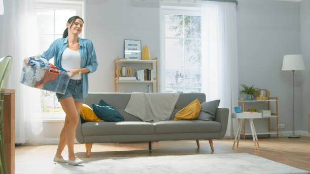 beautiful and happy brunette young woman moving towards the washing machine in homely jeans clothes. she carrying laundry basket to the washer. bright and spacious living room with modern interior. - spring cleaning women cleaning dancing imagens e fotografias de stock