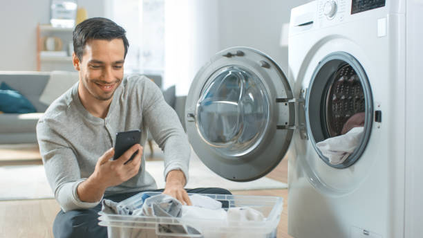 Handsome Smiling Young Man in Grey Jeans and Coat Sits in Front of a Washing Machine and Uses His Smartphone. He Loads Washer with Dirty Laundry. Bright and Spacious Living Room with Modern Interior. Handsome Smiling Young Man in Grey Jeans and Coat Sits in Front of a Washing Machine and Uses His Smartphone. He Loads Washer with Dirty Laundry. Bright and Spacious Living Room with Modern Interior. laundromat laundry residential structure cleaning stock pictures, royalty-free photos & images