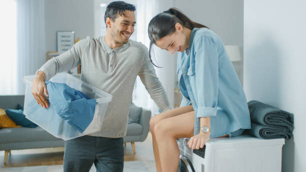 schöne junge paar sprechen in einem wohnzimmer zu hause. mädchen sitzt auf einer waschmaschine, mann hält wäsche. thay sind happy und lachen. zimmer ist modern eingerichtet. - laundry husband housework men stock-fotos und bilder