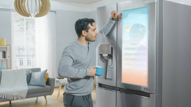 istock Handsome Young Man is Standing Next to a Refrigerator While Drinking His Morning Coffee. He is Checking a To Do List on a Smart Fridge at Home. Kitchen is Bright and Cozy. 1208033035