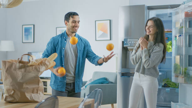 美しい若いカップルは、キッチンで楽しみを持っています。人間はオレンジとジャグリングしています。女の子は拍手、彼のために応援し、彼らは笑っています。モダンなハイテク冷蔵庫(新 - smiling single object photography orange ストックフォトと画像