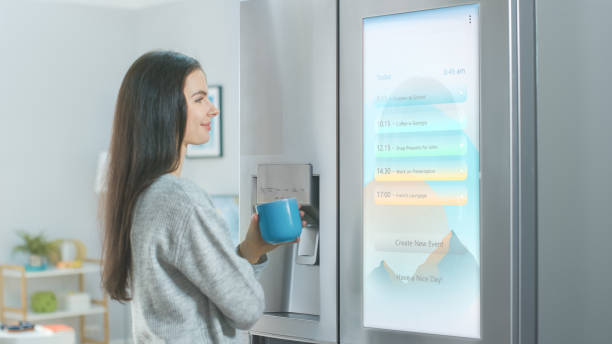 istock Beautiful Young Girl Stands Next to a Refrigerator While Drinking Her Morning Coffee. She is Checking the Weather Forecast and a To Do List on a Smart Fridge at Home. Kitchen is Bright and Cozy. 1208033100