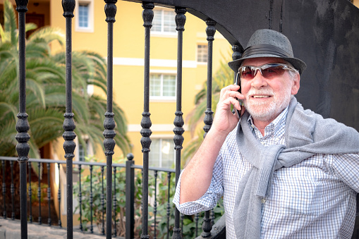 A smiling senior man with beard and white hair talking at the cellphone - standing close to an iron black gate - black cap