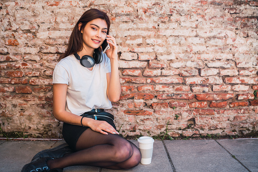 Portrait of young latin woman talking on the phone while sitting outdoors against brick wall. Urban concept.
