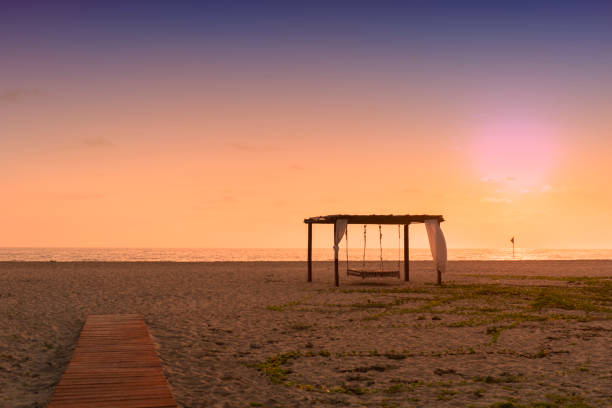 Wooden walkway to the sand beach with hammock cottage decorated with white fabric over sea water and colorful sunset sky background in Baja California, Mexico.
