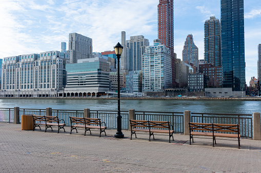 An empty row of benches with a street light on the waterfront of Roosevelt Island along the East River with a view of the Upper East Side skyline in New York City