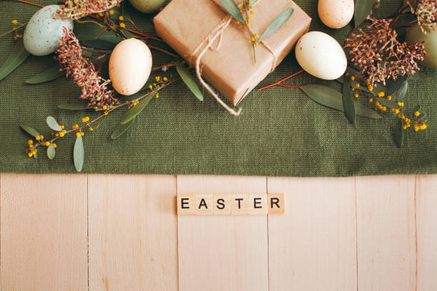 White wooden table with white plate and bowl, pastel colored Easter eggs, fresh garden flowers and brown paper wrapped gift, green table runner