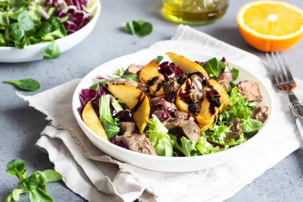 Warm salad with fried chicken liver, caramelized pear, dried cranberries and salad mix with orange dressing on a grey concrete or stone background. Healthy or diet food. Copy space.