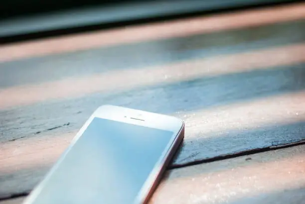 A smart phone with turned-off display placed on a brown wooden table, with sunlight