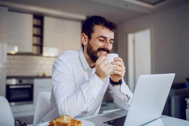 istock Young cheerful smiling attractive bearded man sitting at dinning table, reading an e-mail on laptop and enjoying his fresh morning coffee. 1208102844