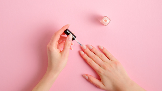 Woman applying polish on nails at home. Female hands with elegant manicure and nail polish bottle on pink background, top view. Beauty treatment and hand care concept